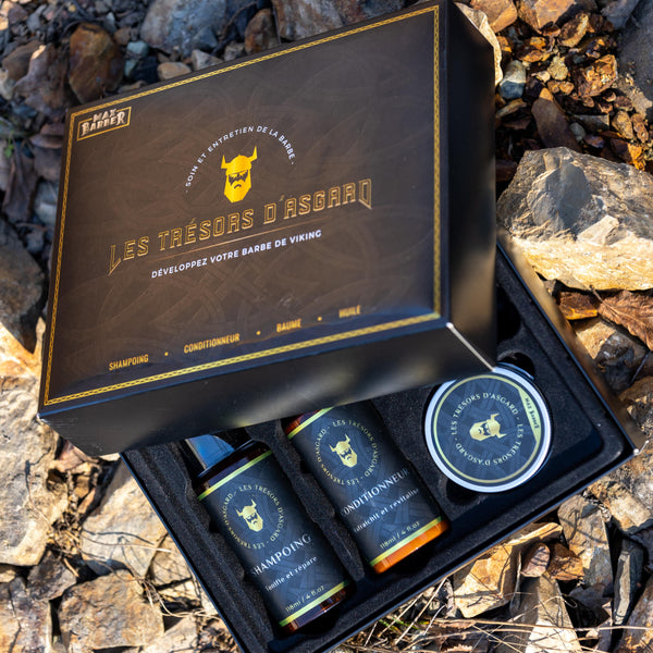 Un coffret de soin pour barbe "Les Trésors d'Asgard" de Max Barber est posé sur un sol rocailleux. Le coffret noir, orné de motifs nordiques et du logo d'un casque viking doré, contient quatre produits : un shampoing, un conditionneur, un baume et une huile. Les flacons et le baume sont élégamment disposés à l'intérieur de la boîte, chaque produit étant étiqueté avec le même design viking. L'emballage robuste et soigné évoque l'univers des guerriers nordiques, soulignant un soin de barbe haut de gamme et ma