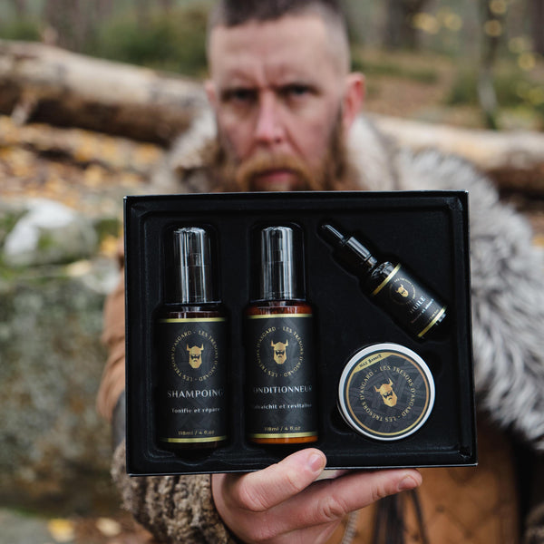 Un homme barbu en arrière-plan, vêtu d'une tenue évoquant les Vikings, présente un coffret de soin pour barbe "Les Trésors d'Asgard" de Max Barber. Le coffret, ouvert au premier plan, contient quatre produits : un shampoing (118 ml), un conditionneur (118 ml), une huile pour barbe (30 ml), et un baume. Chaque flacon et pot arbore un design noir et doré avec un logo de casque viking. L'homme, dans un environnement extérieur boisé, met en valeur le côté robuste et authentique de la gamme, destinée à ceux qui 